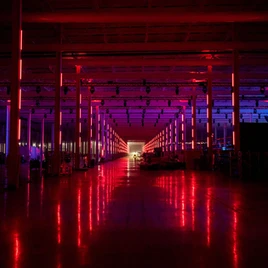 Intérieur d'un grand entrepôt ou hall industriel éclairé par des lumières rouges et violettes, créant un effet de perspective avec des colonnes et un sol réfléchissant